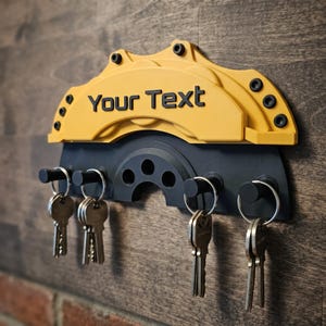 Peut inclure: Un porte-clés mural jaune et noir en forme d'étrier et de disque de frein de voiture. La section jaune de l'étrier porte le texte noir Your Text. Quatre crochets noirs dépassent de la base grise foncée, chacun portant un anneau métallique.