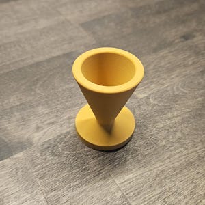 May include: A matte yellow egg cup with a conical top section and a circular base. The object features a smooth, uniform finish and a wide, open rim designed to hold a single egg securely in an upright position for serving or display purposes.