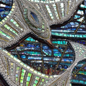 May include: Close-up of a mosaic artwork featuring a pattern of blue, green, and silver iridescent glass tiles. The mosaic is made up of small, irregular pieces of glass that are arranged in a geometric pattern.