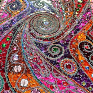 May include: A colourful mosaic table top with a swirling pattern of red, orange, purple, green, and blue glass tiles. The table top is made of small, irregular pieces of glass that are arranged in a geometric pattern.