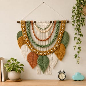 Puede incluir: Un tapiz de macramé colgado de una vara de madera oscura, con arcos superpuestos de cuerda en color crema, verde salvia y terracota. El diseño incluye cuentas de madera, patrones trenzados intrincados y varios elementos de flecos en forma de hoja.