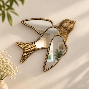 Peut inclure: Un miroir mural en forme d'oiseau en plein vol avec un cadre texturé de couleur or antique. Les ailes et le corps de l'oiseau comportent des panneaux de verre réfléchissants insérés dans la structure métallique, ornée de motifs de plumes gravés.