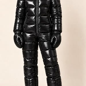 Puede incluir: Un traje de nieve negro brillante con acabado de alto brillo, cremallera frontal completa, capucha y acolchado horizontal. El conjunto incluye manoplas negras a juego y botas negras, creando un aspecto aislado y uniforme.