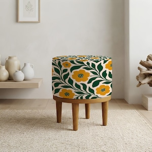 Puede incluir: Un taburete redondo con tapicería de tela color crema con un patrón repetitivo de flores amarillo mostaza y hojas verde oscuro. La pieza se apoya sobre cuatro patas de madera cortas y cilíndricas con un acabado natural marrón claro.