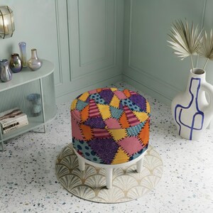 May include: A round ottoman featuring a colorful patchwork pattern with various geometric shapes, swirls, and polka dots in shades of orange, yellow, pink, purple, and blue, supported by four short, sturdy white legs on a circular patterned rug.