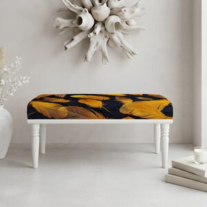 May include: A rectangular bench featuring a dark navy blue upholstered seat with a repeating pattern of golden yellow feathers. The bench is supported by four white, turned wooden legs that provide a stable base for the cushioned seat.