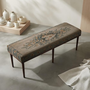 May include: A rectangular upholstered bench with four dark brown tapered wooden legs. The seat features a muted gray fabric with a central heart-shaped motif surrounded by floral patterns in shades of blue, orange, and green, with striped sides.