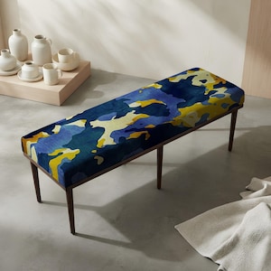 May include: A rectangular bench featuring a cushioned seat upholstered in a vibrant abstract pattern of deep blue, light blue, and yellow shapes. The bench is supported by four slender, dark brown wooden legs that taper toward the floor.