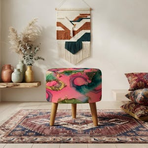 May include: A round ottoman featuring a vibrant abstract pattern with swirls of hot pink, teal, and gold tones. The piece is supported by three light-colored wooden legs, providing a stable base for the circular, cushioned seat top.