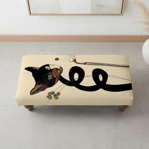 Puede incluir: Un otomán rectangular con una superficie de color crema que presenta una ilustración estilizada de un gato negro. El gato tiene una cola larga y enroscada, con patrones coloridos y complejos en la cabeza y el cuello. Se apoya en cuatro patas de madera.