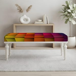 May include: A rectangular bench featuring four white turned legs and a cushioned seat upholstered in a vibrant geometric pattern. The fabric displays a gradient of colors transitioning from yellow and orange on the left to red and purple on the right.