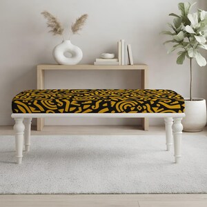 May include: A rectangular bench featuring a seat upholstered in a black fabric with a repeating mustard yellow abstract pattern. The bench is supported by four white turned wooden legs that provide a stable base for the cushioned seating area.