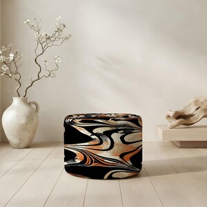 May include: A cylindrical ottoman featuring a bold, abstract marbled pattern in shades of black, white, and burnt orange. The fabric surface displays fluid, swirling lines that create a dynamic visual effect across the entire rounded exterior.