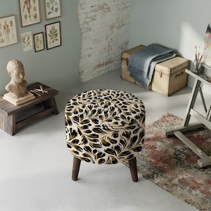 May include: A round ottoman featuring a cream-colored fabric base with a repeating pattern of black and gold leaf-like shapes. The piece is supported by four dark brown wooden legs that taper slightly toward the floor, providing a stable base.