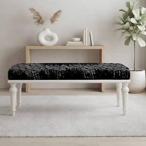 May include: A rectangular bench featuring a black upholstered seat with a white intricate geometric pattern. The bench is supported by four white wooden legs with a turned, spindle-style design, providing a stable base for the cushioned top.