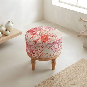 May include: A round ottoman featuring a cream-coloured fabric top with a vibrant red and pink floral line-art pattern. The seat is supported by four short, turned wooden legs in a light natural finish, providing a stable base for the circular cushion.