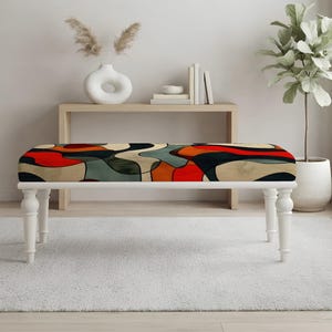 May include: A rectangular bench featuring a cushioned seat with an abstract pattern of wavy, interlocking shapes in shades of red, black, beige, and muted green. The bench is supported by four white, turned wooden legs with a classic spindle design.
