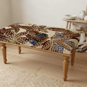 May include: A rectangular upholstered bench featuring four light-toned wooden legs with a turned design. The seat cushion is covered in a fabric displaying a large-scale floral pattern in shades of brown, cream, and blue with intricate dot details.