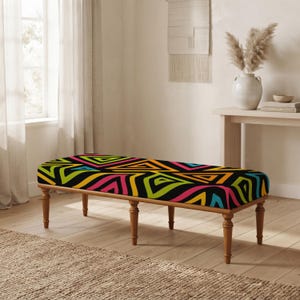 May include: A rectangular bench featuring a vibrant, multi-colored geometric pattern of triangles in shades of neon green, hot pink, orange, and blue on a black background, supported by six turned wooden legs with a natural light brown finish.