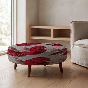 Puede incluir: Un otomán redondo con una base de tela gris y un patrón repetitivo de grandes hojas estilizadas de color rojo intenso. La pieza se apoya sobre cuatro patas cortas de madera cónica en un acabado marrón medio, ofreciendo una base estable.