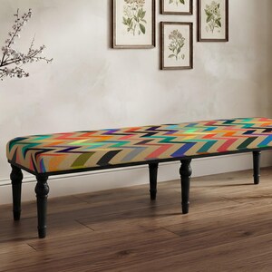 May include: A long rectangular bench featuring a vibrant, multi-colored chevron pattern in shades of orange, blue, green, and yellow. The bench is supported by four dark, turned wooden legs that provide a sturdy base for the upholstered seat.