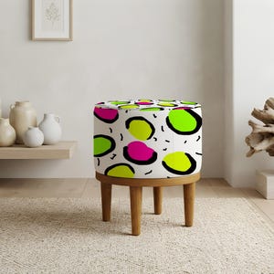May include: A round ottoman featuring a white fabric upholstery with a repeating pattern of black-outlined neon green and hot pink abstract shapes, supported by four short, light brown wooden legs in a cylindrical, low-profile design.