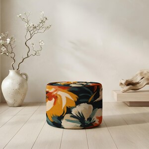 May include: A cylindrical ottoman featuring a bold, abstract floral pattern with large petals in shades of deep navy blue, vibrant orange, golden yellow, and soft cream. The fabric has a smooth texture and covers the entire round surface of the piece.