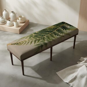 May include: A rectangular bench with a taupe upholstered seat featuring a large green fern leaf print. The bench has four dark brown wooden legs. The seat is flat and elongated, providing a surface for seating, while the legs are slender and straight.