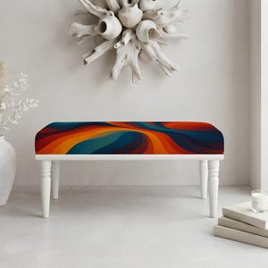 May include: A rectangular bench featuring a vibrant, multi-colored fabric seat with a swirling, wave-like pattern in shades of deep blue, orange, yellow, and red. The bench is supported by four white, turned wooden legs with a smooth finish.