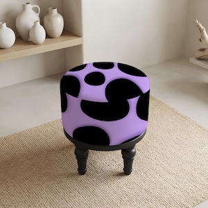 May include: A round ottoman featuring a light purple fabric upholstery with a bold black cow-print pattern. The seat is supported by four short, turned black wooden legs that provide a stable base for the circular cushioned top.