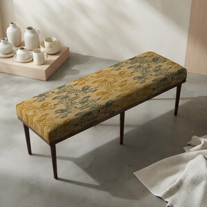 May include: A rectangular bench with four dark brown wooden legs and a cushioned seat upholstered in a mustard yellow fabric featuring a dark green botanical pattern of leaves and birds, positioned in a room with light-colored walls and flooring.