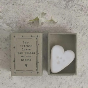 May include: A light grey cardboard box with a lid featuring the black printed text: Best friends leave paw prints on our hearts, with a small heart icon. Inside the open box sits a smooth, white, heart-shaped stone with a paw print indentation.