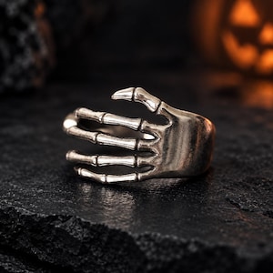 May include: A silver-toned metal ring shaped like a skeletal hand. The design features articulated finger bones that wrap around the band, with the fingers extending to form the main structure of the ring. The finish has a dark, weathered appearance.