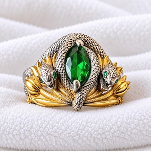 Può includere: Un anello con una pietra centrale verde smeraldo taglio marquise circondata da un serpente in argento testurizzato. La fascia è decorata con fogliame dorato e due teste di serpente laterali, ciascuna con piccoli occhi in pietra verde.
