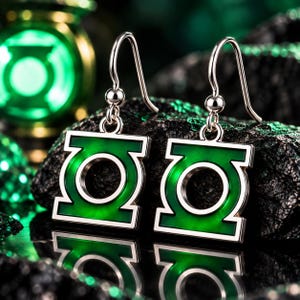 以下が含まれることがあります： グリーンランタンのロゴをあしらったシルバー調のドロップピアス。各ピアスは正方形のフレームと円形の中心部で構成され、鮮やかなグリーンのエナメルが施されています。シンプルなシルバー調の金属製フックから吊り下げられています。