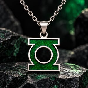 May include: A silver-toned metal pendant featuring the Green Lantern symbol, characterised by a square frame with a central circular cutout. The recessed areas of the pendant are filled with a textured, vibrant green inlay, suspended from a silver chain.