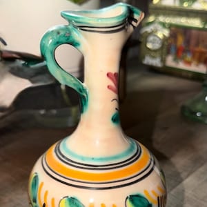 May include: A small ceramic pitcher with a cream-colored base, featuring hand-painted floral motifs in green, yellow, and red. The vessel has a narrow neck, a single curved handle, and horizontal black line accents around the body and rim.