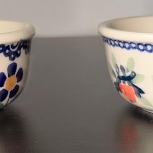 May include: Two small ceramic bowls with a cream-colored base. The left bowl features blue flowers with yellow centers and green leaves. The right bowl displays a pattern of orange fruits with dark blue leaves and a blue decorative border along the rim.