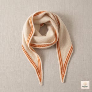 May include: A triangular scarf in a soft cream color featuring two parallel orange stripes along the edges. The scarf has a pointed shape and a smooth, knit-like texture. A small white square label in the corner displays the text FEMME.