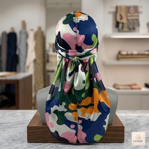 Op de afbeelding: Een satijnen durag met een meerkleurig camouflagepatroon in tinten donkerblauw, bosgroen, feloranje, lichtroze en wit. Het hoofddeksel is aan de achterkant gestrikt met lange stoffen uiteinden en toont onderaan de tekst FEMME.