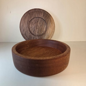 May include: A round, dark brown wooden bowl with a smooth, polished finish and visible wood grain. Behind the bowl, a matching circular wooden lid is positioned upright, featuring a recessed centre circle and a flat, circular outer rim.