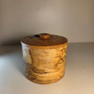 May include: A cylindrical wooden container with a matching lid featuring a small central knob. The exterior displays a natural light tan wood grain with dark, irregular swirling patterns and knots throughout the surface of the vessel.