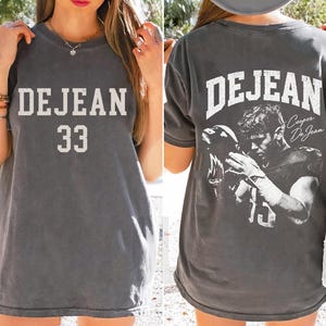 Pode incluir: Uma t-shirt de manga curta cinzento-carvão com o texto DEJEAN e o número 33 na frente. As costas exibem o texto DEJEAN, um gráfico de um jogador de futebol americano segurando um capacete e a assinatura Cooper DeJean em tinta branca.