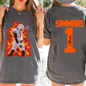 Könnte beinhalten: Ein anthrazitfarbenes T-Shirt mit einem Frontaufdruck eines Footballspielers in weißer Uniform, umgeben von orangefarbenen Flammen und dem Text #1. Auf der Rückseite steht der Name SIMMONS in Orange über einer großen orangefarbenen Nummer 1.