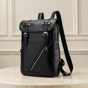 Puede incluir: Una mochila negra con solapa, dos correas verticales y hebillas de metal plateado. La parte frontal incluye un bolsillo diagonal con cremallera plateada. La mochila cuenta con correas de hombro negras ajustables y un acabado liso.