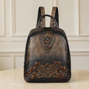 May include: A dark brown leather backpack featuring a two-tone gradient finish. The bag is adorned with intricate embossed floral patterns on the front flap and lower section. It includes a top handle and two shoulder straps for carrying.