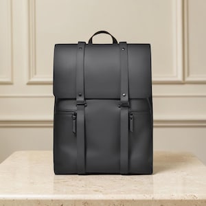 Puede incluir: Una mochila negra mate con cierre de solapa, dos correas verticales y botones de presión metálicos. La bolsa incluye un asa superior, dos bolsillos frontales con cremallera con tiradores negros a juego y un diseño exterior liso y limpio.