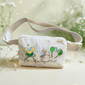 May include: A cream-coloured belt bag featuring a graphic of Tinker Bell, a pair of green glittery mouse ears, and a yellow and green bow. The text "Fly with faith and trust" is printed in black script below the illustrations, with an adjustable strap.
