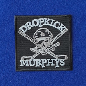 May include: A square black embroidered patch featuring a white graphic of a skull wearing a hockey helmet, positioned in front of two crossed hockey sticks. The text DROPKICK is arched above the skull, and MURPHYS is printed below.