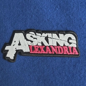 Puede incluir: Un parche bordado que presenta el texto Asking Alexandria. La palabra Asking es de color blanco con una textura desgastada, y la palabra Alexandria es de color rosa. Todo el diseño está delineado con hilo negro sobre un fondo azul sólido.